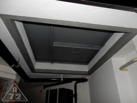 Pohled ze spodního patra na uzavřený průlez do bojového patra třídílným poklopem, tedy tak, jak to tenkrát mělo být. <br>Vzadu je vidět nádrž v umývárně a rozvod vody, který kopíruje tvar průlezu kvůli tomu, aby nepřekážel při sestupu do podzemí.